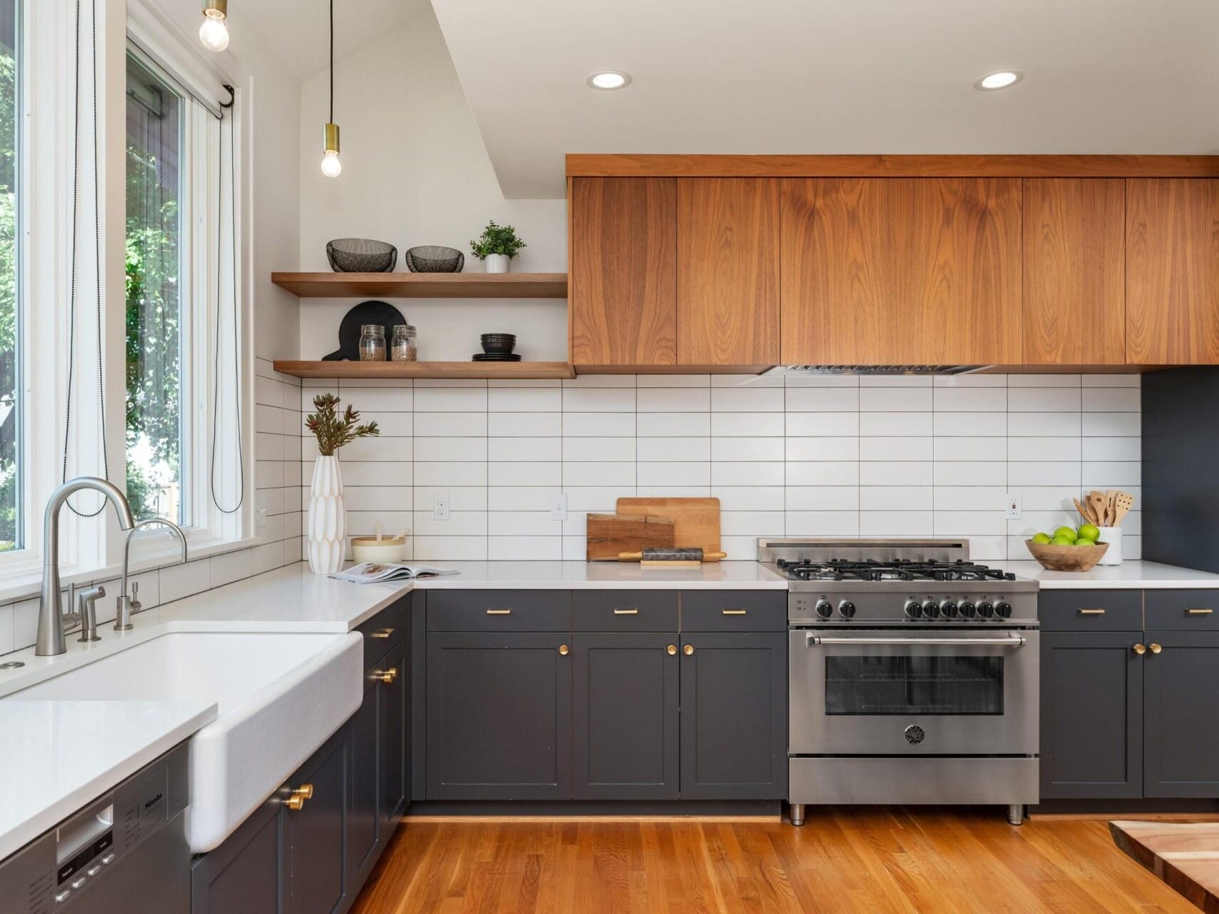 Modern kitchen with dark cabinets, wooden countertops, and a white tile backsplash. A stainless steel stove and range hood are centered. Sunlight streams through a large window by a white farmhouse sink. Accessories include bowls and a fruit basket.