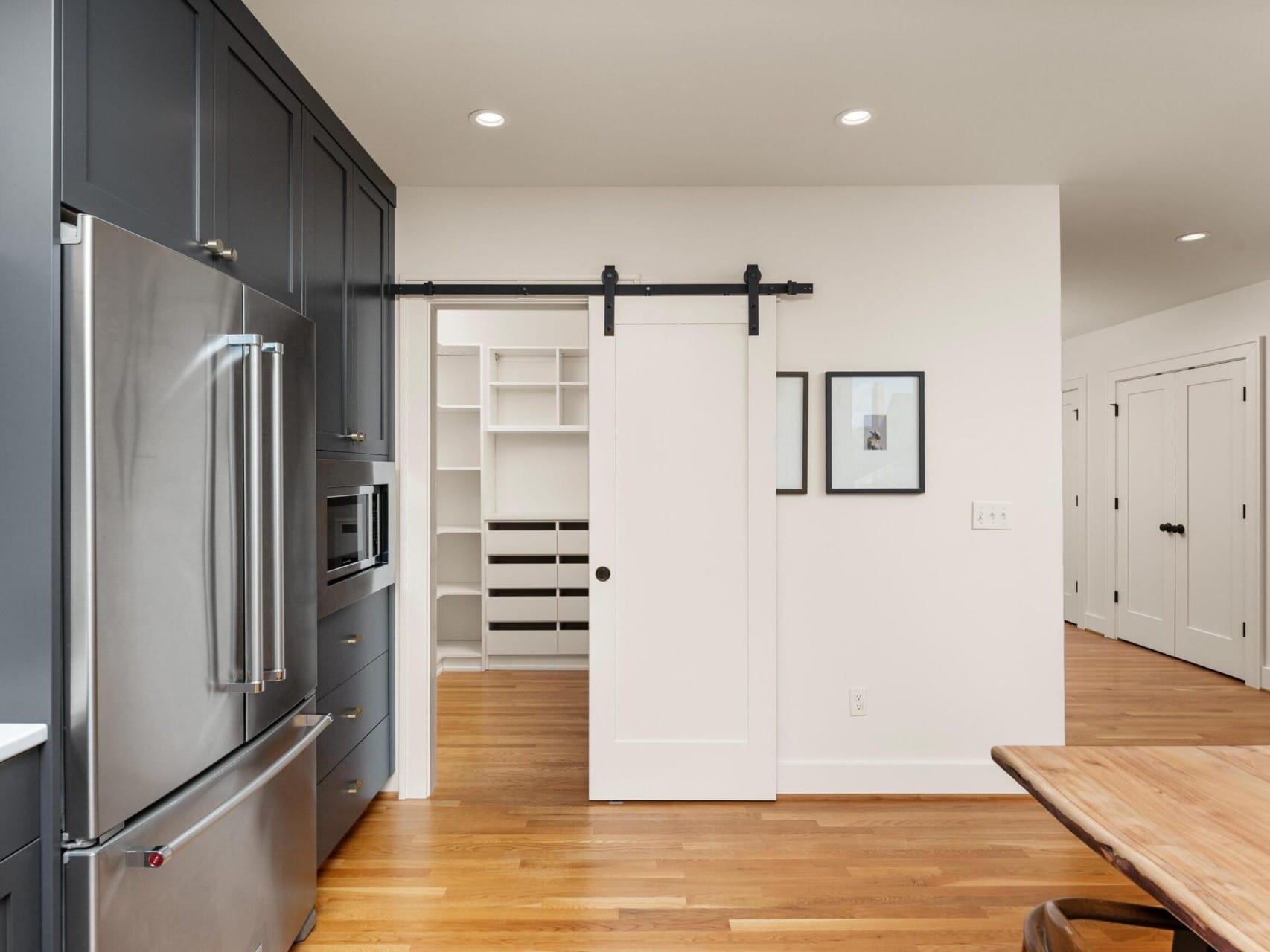 Modern kitchen with stainless steel refrigerator, dark cabinets, and light wood flooring. A sliding barn door leads to a pantry with white shelves. Wooden dining table visible on the right.