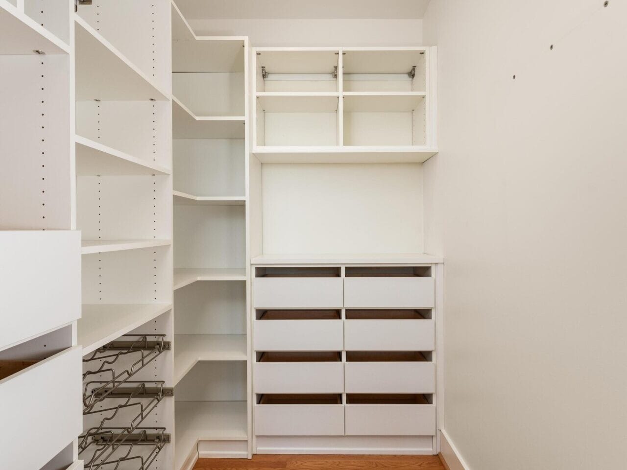 A spacious, empty walk-in closet with wooden flooring and white shelving. It features multiple open shelves, drawers, and hanging racks, all ready for organizing clothes and accessories. A ceiling light illuminates the space.