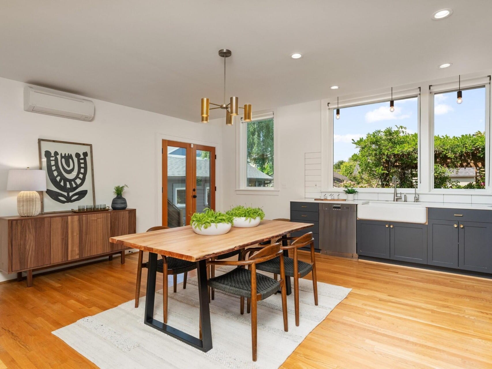 A modern dining area with a wooden table and six chairs on a light rug. Large windows with a view of greenery, a sideboard with a decorative lamp, and pendant lighting complete the room. The floor is polished wood, and the kitchen area is visible.