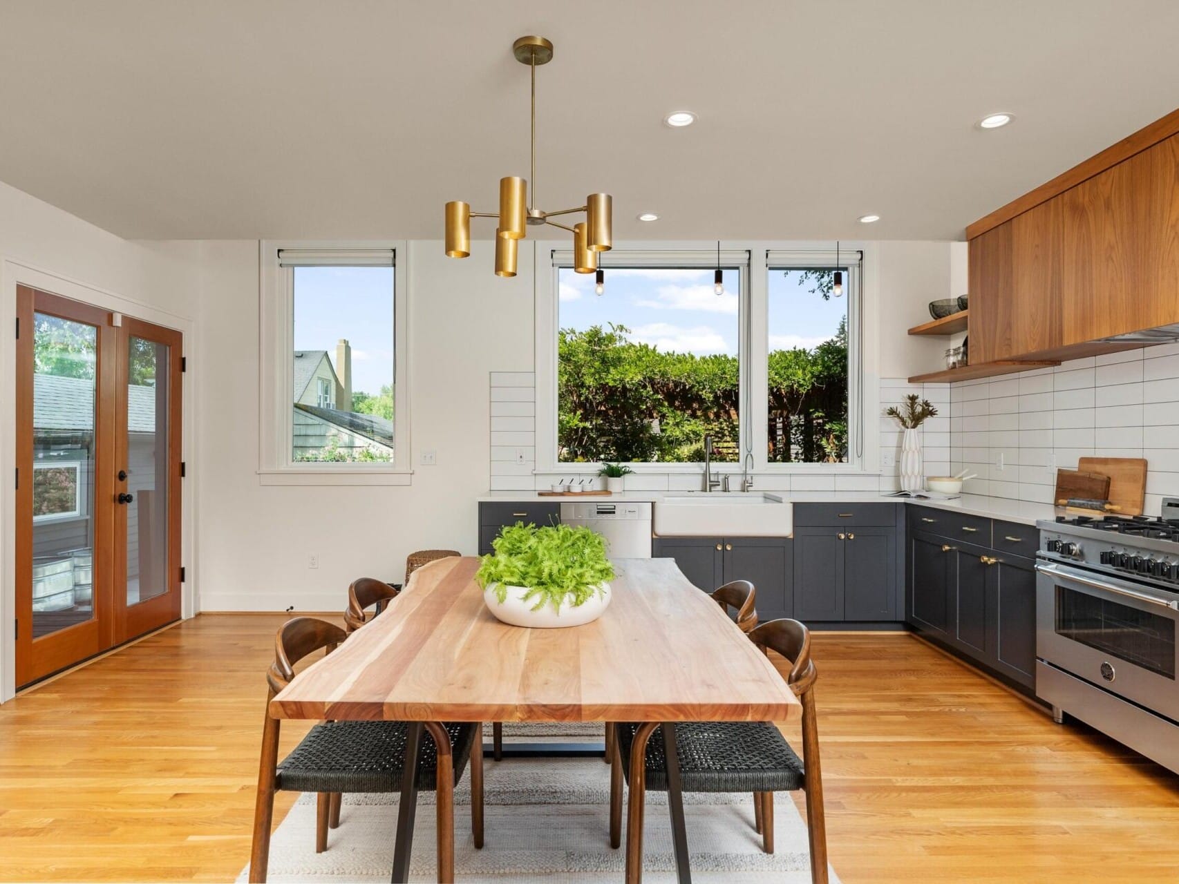 Modern kitchen with wooden dining table and six chairs, a hanging gold light fixture, and a potted plant centerpiece. The kitchen features dark cabinets, white tile backsplash, stainless steel appliances, and large windows with outdoor views.