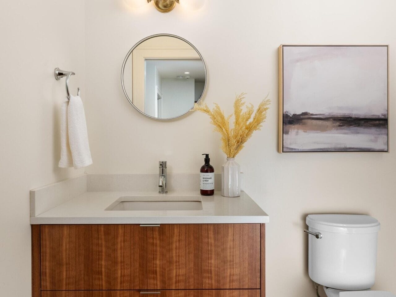 A modern bathroom features a wooden floating vanity with a round mirror above it, illuminated by a wall sconce. A white towel hangs on a rack, and a potted plant and soap dispenser sit on the counter. A toilet is positioned next to a piece of abstract art.