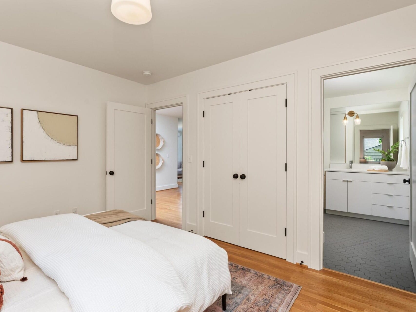 Bedroom with a bed on a rug, adorned with white and brown pillows. Two abstract wall art pieces hang above. The room features wooden flooring, two closed closet doors, and an open door leading to a bathroom with hexagonal tiles and a white vanity.