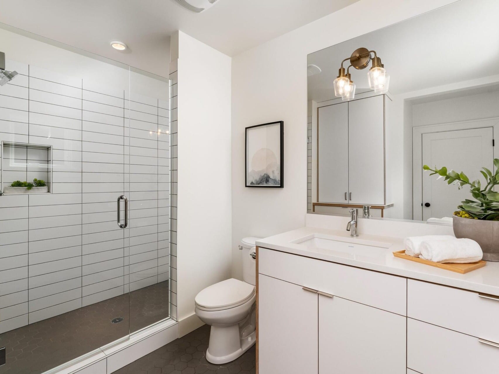 A modern bathroom featuring a glass-enclosed shower with white tiled walls, a sleek white vanity with a large mirror, a white toilet, and a potted plant for decoration. A small framed artwork hangs on the wall. Bright, well-lit space.