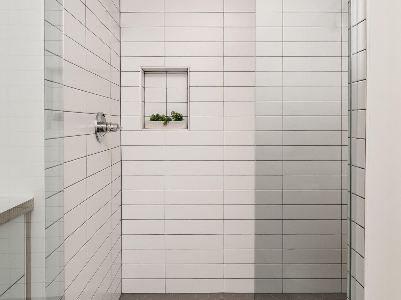 A modern shower with white horizontal subway tiles and a glass door. The shower features a silver showerhead and a small built-in niche with decorative greenery. The floor is covered in hexagonal gray tiles.