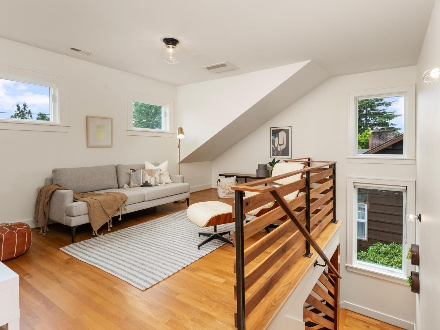 A modern, airy loft space with a gray sofa and light-colored rug on hardwood floors. Three windows allow natural light. A wooden staircase railing leads down, and a stylish white chair adds an accent. A pouf and decor items complete the look.