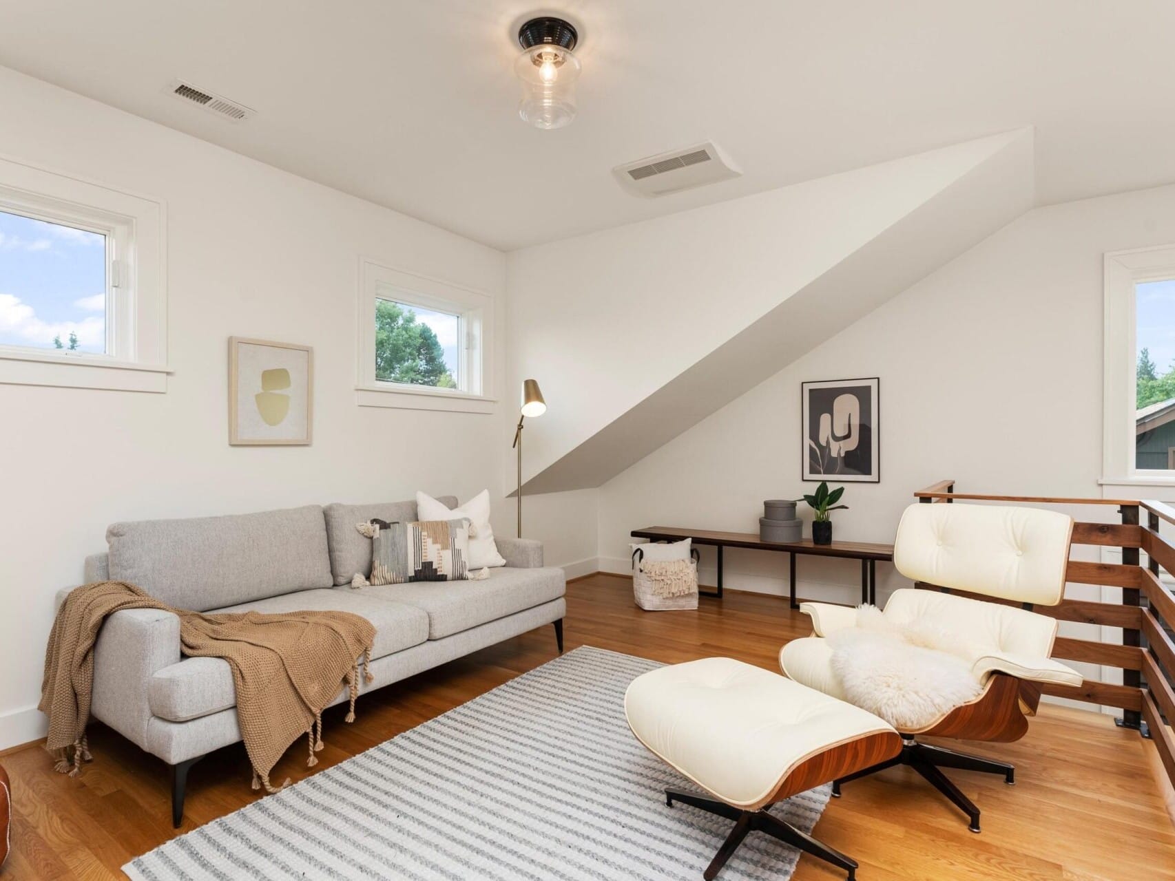 A modern living room with a gray couch, white cushions, and a tan throw blanket. A striped rug covers the wooden floor. A white lounge chair and matching ottoman sit nearby. Art pieces hang on white walls, and a large window brings in natural light.