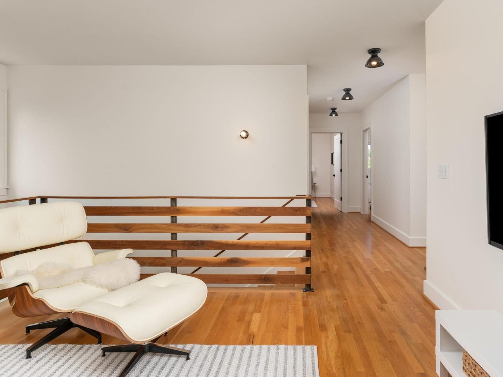 A modern living space with a white lounge chair and ottoman on a light-colored rug. The room features wooden floors and a wooden railing. A hallway leads to other rooms, and a wall-mounted TV is partially visible on the right.