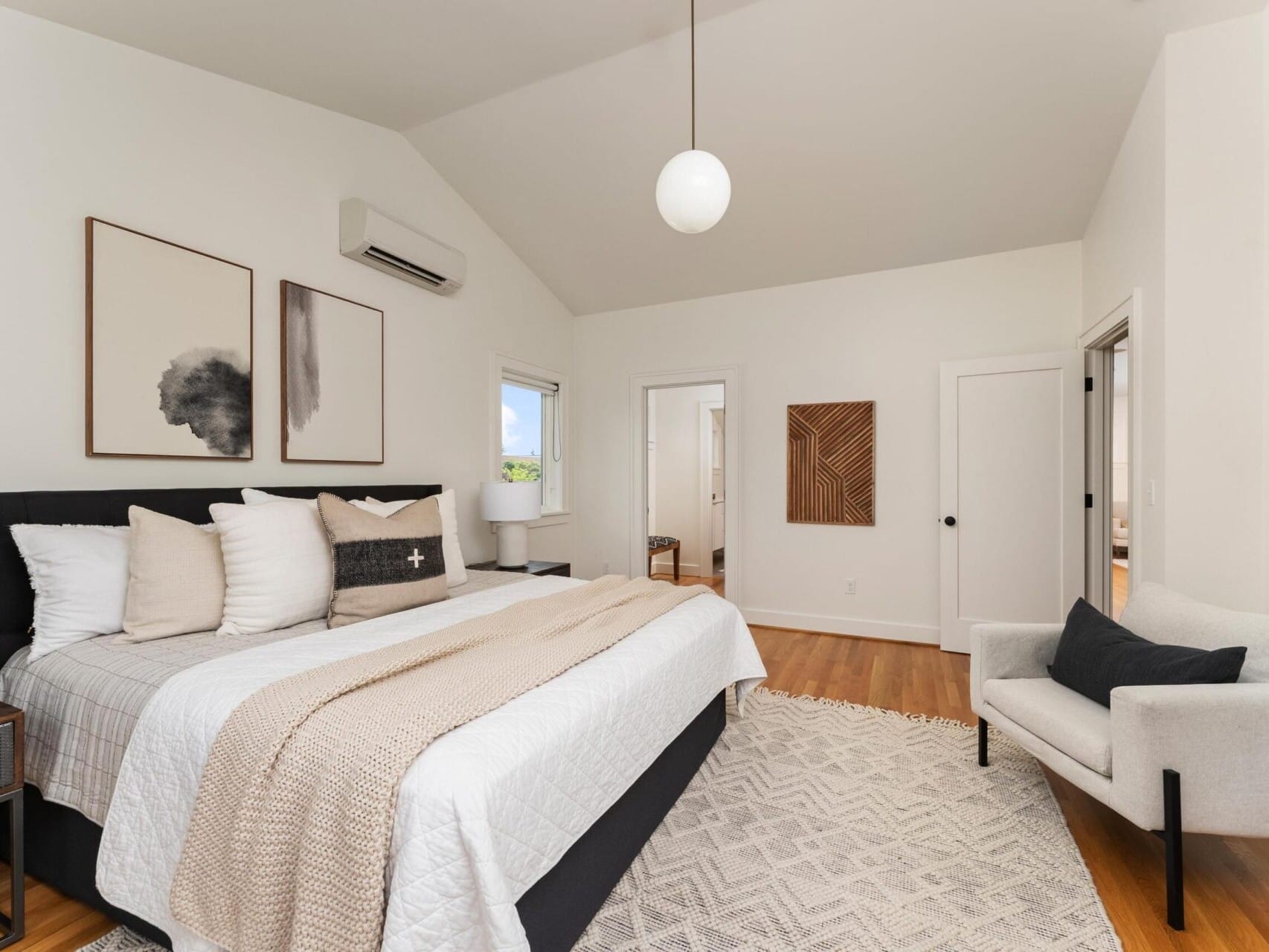 A modern bedroom with a sloped ceiling features a neatly made bed with white and beige bedding. Two abstract paintings hang above the bed. A gray armchair and a small lamp complete the space, which is brightened by natural light from a window.