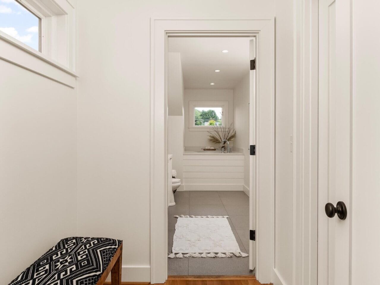 A hallway with wooden floors leads to a bright bathroom. A bench with a black and white geometric cushion is on the left. The bathroom features a white rug, a wide sink, and a decorative plant by a window.