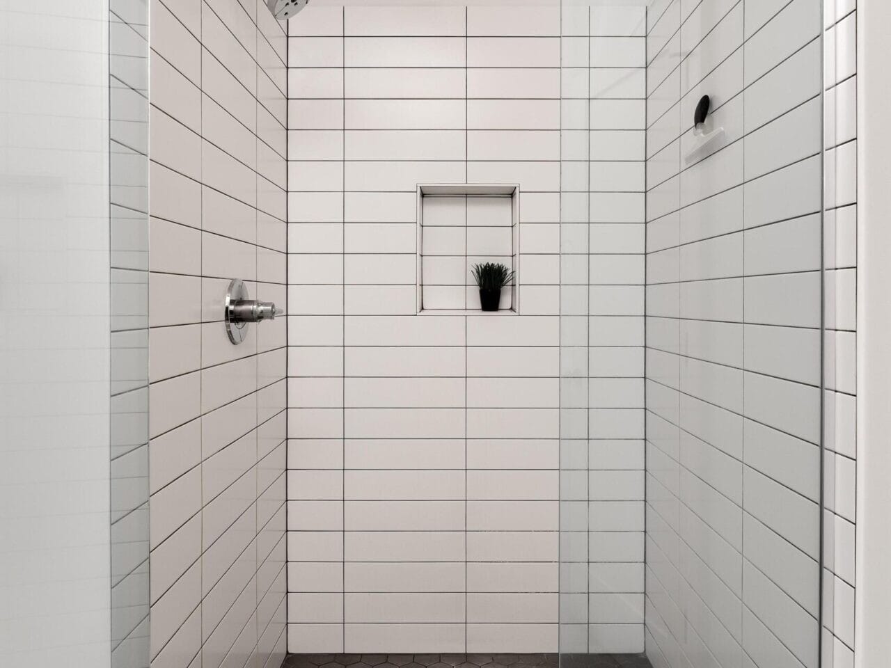 A modern shower with white horizontal tile walls and a glass door. The floor has dark hexagonal tiles, and theres a recessed shelf holding a small plant. A metal showerhead and control are visible on the wall.