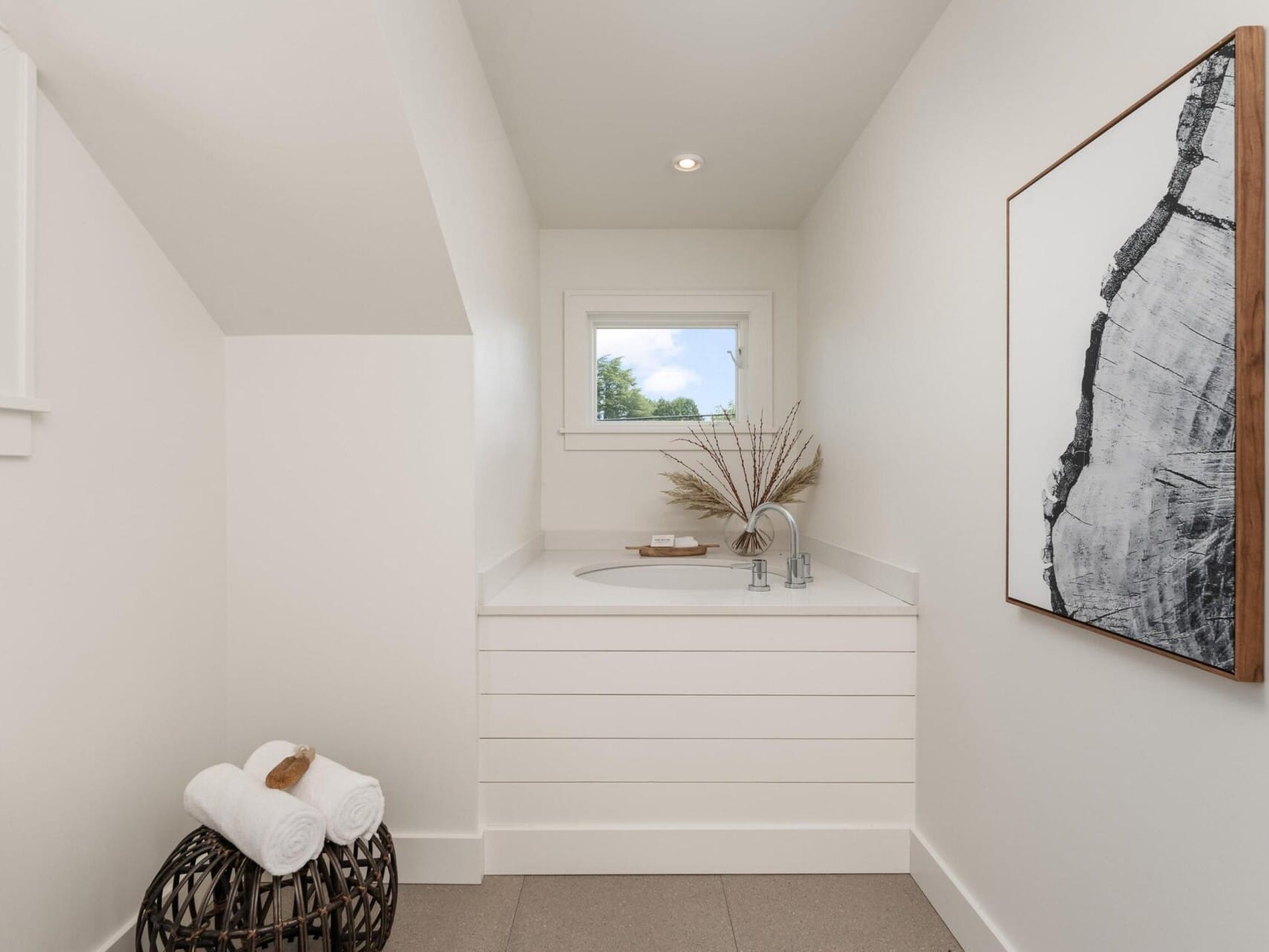 A minimalist bathroom with a small white vanity, a round mirror, and a window above. A vase with dried plants sits on the counter. Rolled towels in a wicker basket are on the floor, and a large abstract artwork hangs on the wall.