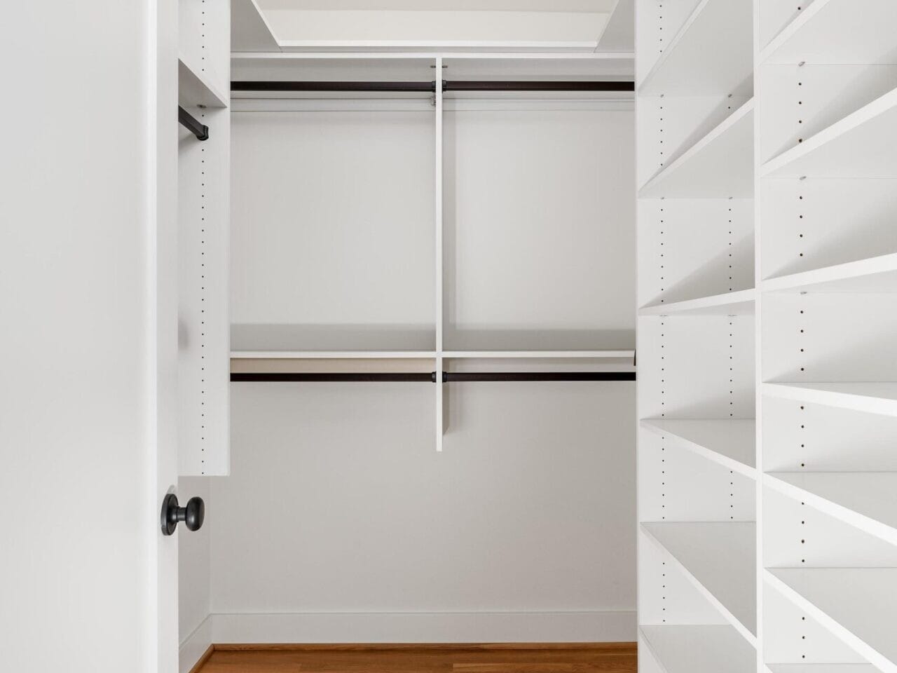 A walk-in closet with white shelves and rods for hanging clothes. The floor is made of light brown hardwood, and theres a recessed light in the ceiling. The closet door is partly visible on the left.