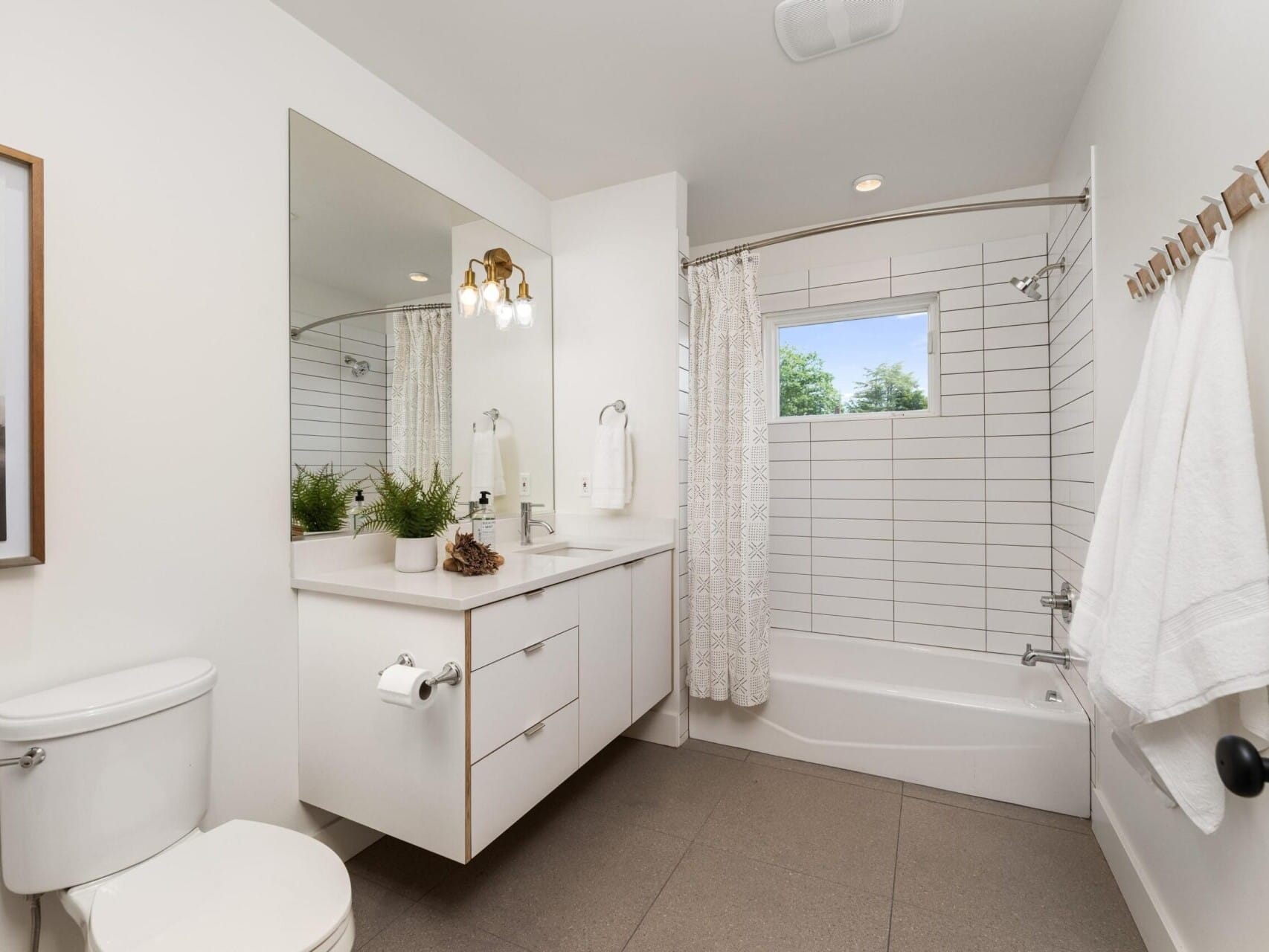 A modern bathroom with a white vanity, large mirror, and overhanging lights. Theres a toilet on the left and a shower-tub combo with white tiles on the right. A small window and towel rack enhance the minimalist decor. A framed picture is on the wall.