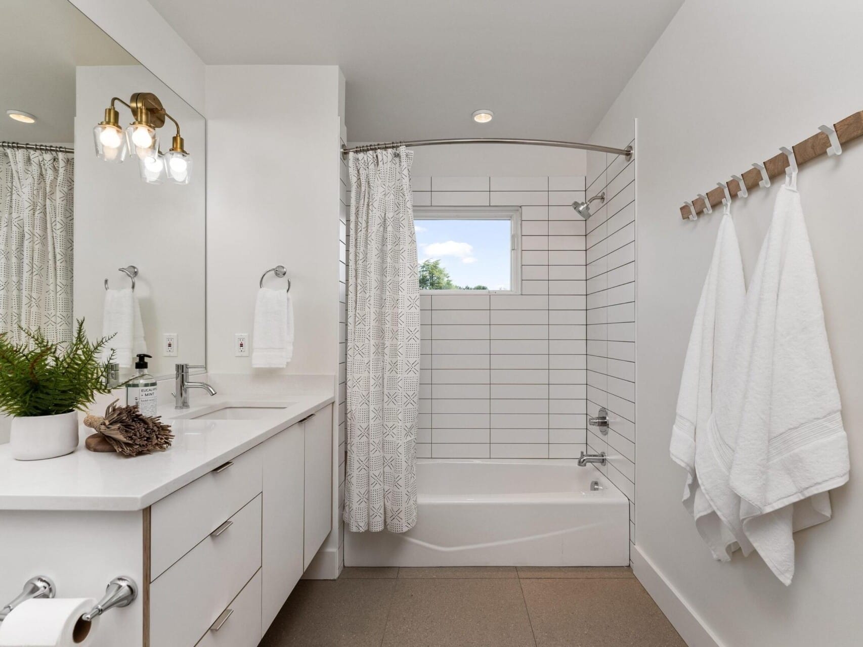 Modern bathroom with a white vanity, double sinks, and a large mirror. A potted plant and rolled towels are on the counter. The room features a bathtub with a white shower curtain, white tiled walls, and towels hanging on wooden hooks.