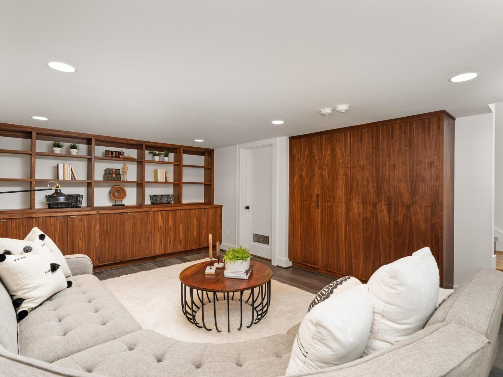 A cozy living room with a gray sectional sofa, white and black cushions, and a round wooden coffee table. The background features a wall of wooden bookshelves with decorative items and a large wooden cabinet. Stairs lead up to the right.