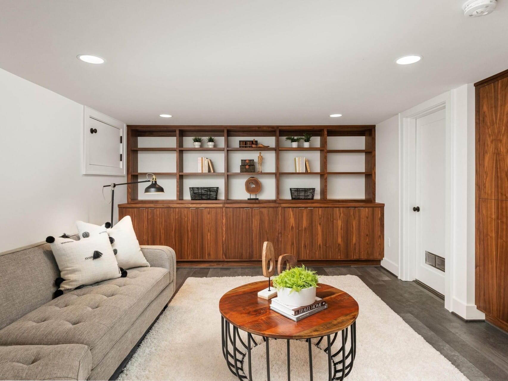 A cozy living room with a gray sofa adorned with cushions, a round wooden coffee table with green plants, a cream rug, and a built-in wooden bookshelf lining the back wall. The space is well-lit with recessed lighting.