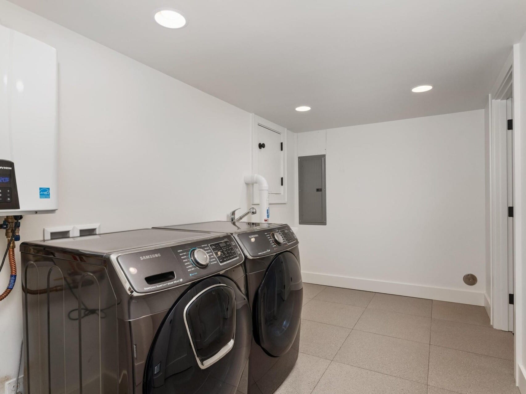 A modern laundry room with two dark-colored front-load washing machines. The room has a white water heater mounted on the wall, recessed lighting, and a tiled gray floor. Walls and ceiling are white, creating a bright, clean space.