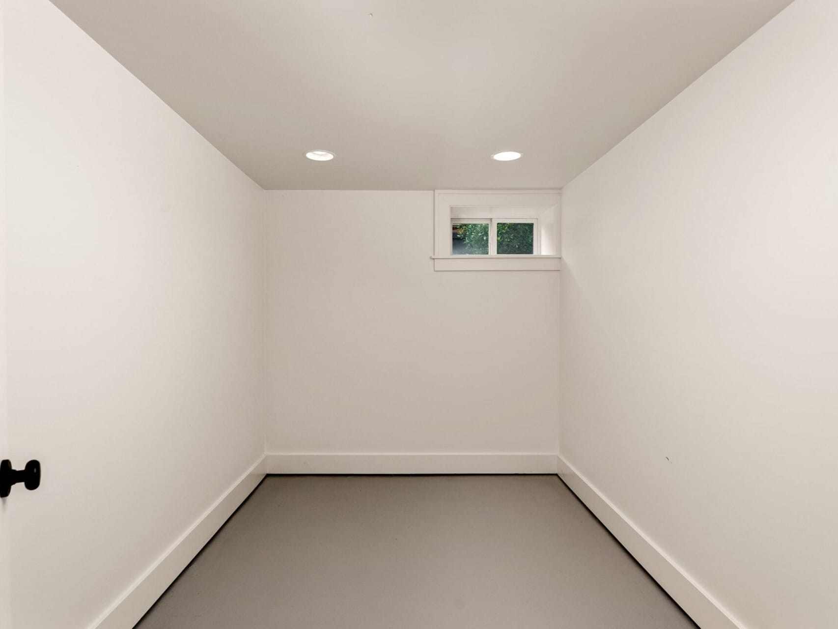 A small, empty room with white walls, a light gray floor, and a single small window high on the back wall. The ceiling has recessed lighting, and theres a black door handle on the left.