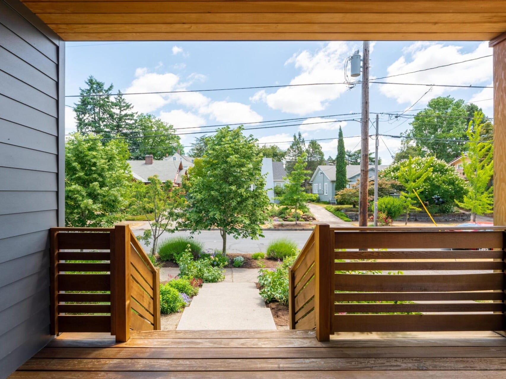 View from a wooden porch overlooking a suburban street with lush greenery and trees. The sky is bright and partly cloudy, and small houses are visible in the background. A pathway leads from the porch steps to the sidewalk.