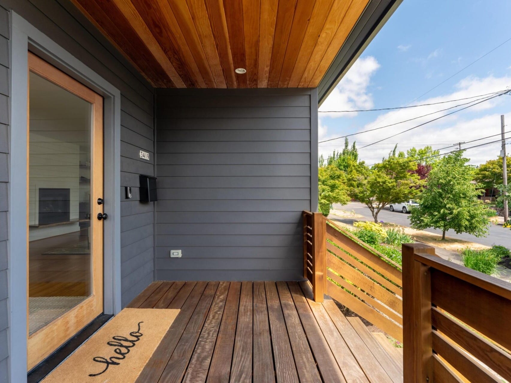 A modern wooden porch with horizontal paneling and a ceiling overhang. A door with a glass window has a hello mat in front. The railing is wooden, and surrounding trees are visible against a blue sky and a street.