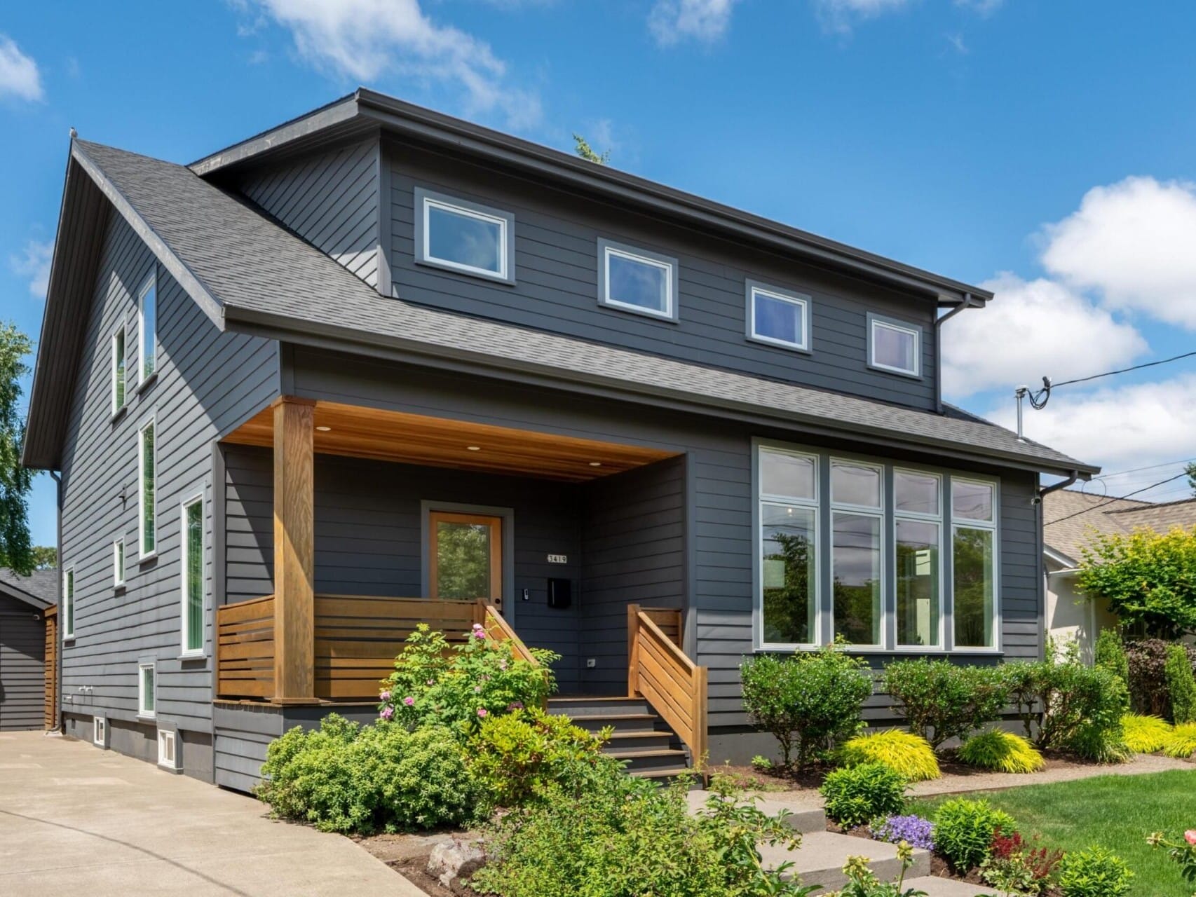 A modern two-story house with dark gray siding and a wooden porch. It features large windows and a well-maintained garden with colorful shrubs and flowers. The sky is clear with a few clouds.