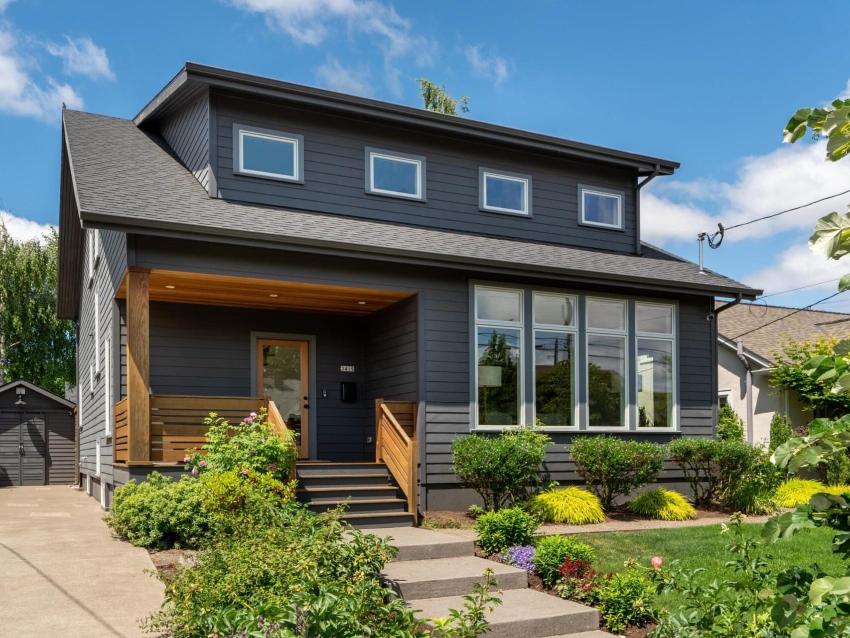 A modern two-story house with dark gray siding, large windows, and a wooden porch. The front yard features neatly trimmed shrubs and colorful flowers, with a clear blue sky overhead. A small driveway leads to a detached garage.