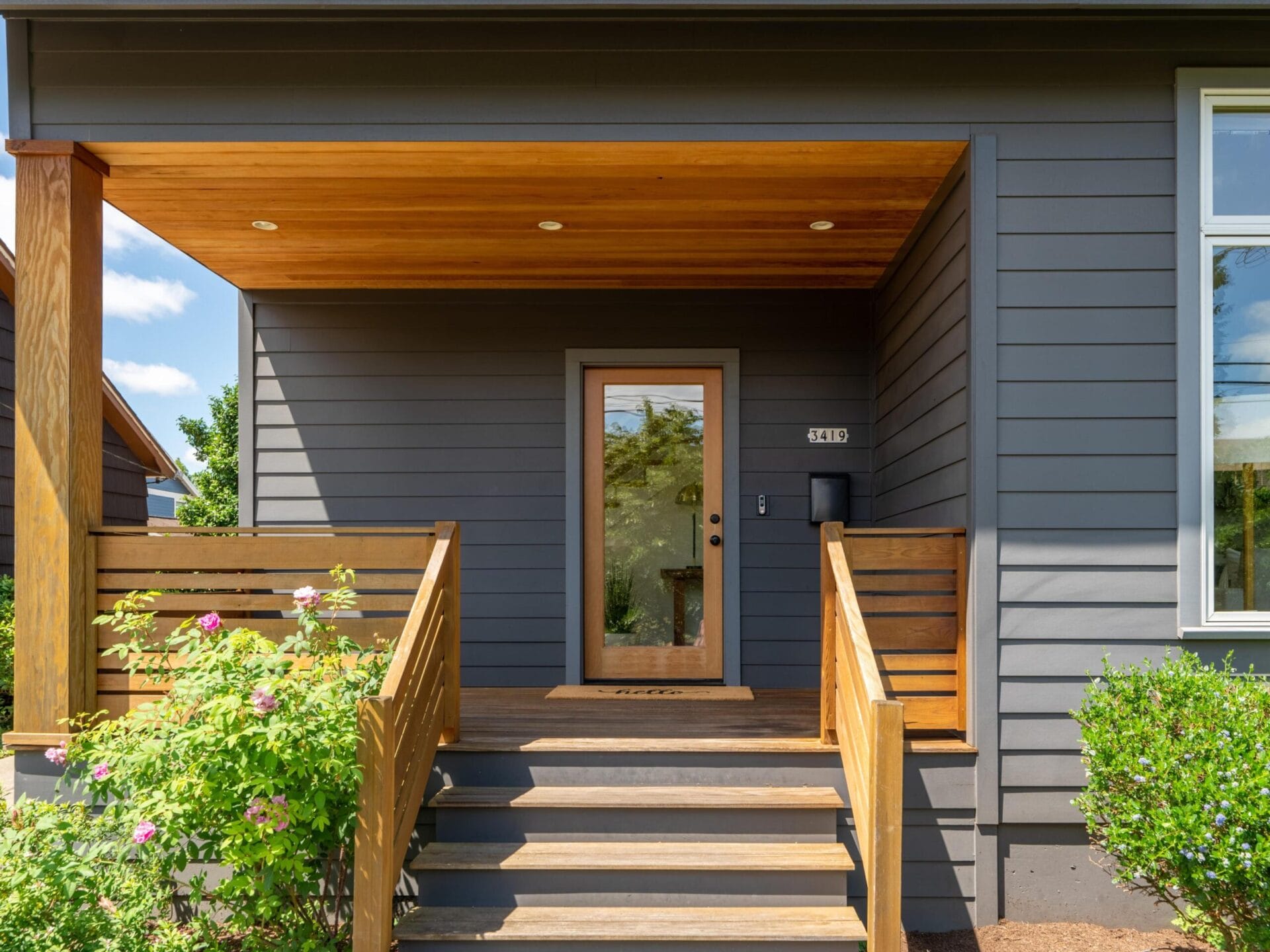 A modern house entrance with a wooden porch and stairs. The exterior features dark gray siding, a glass front door flanked by windows, and a neatly landscaped yard with green shrubs and pink flowers.