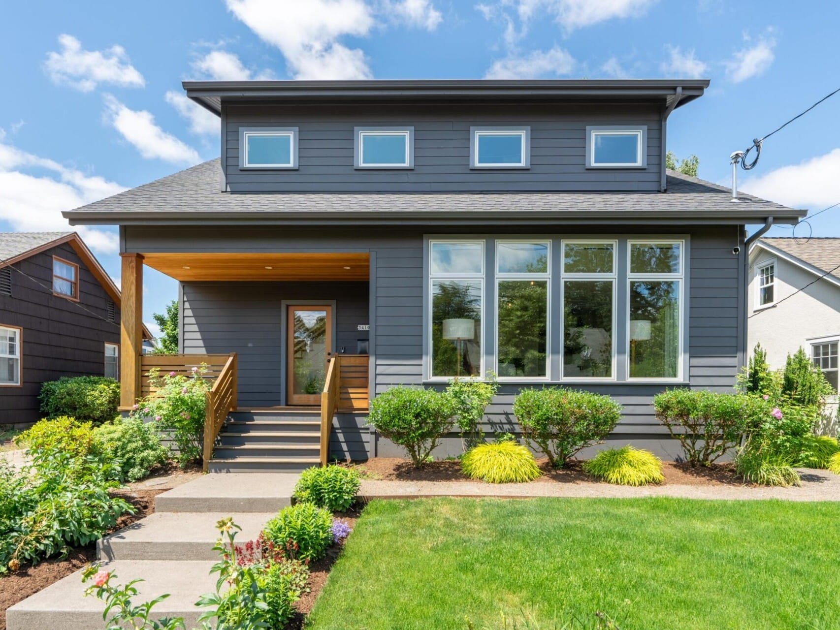 A modern gray house with a wooden porch, large windows, and a well-maintained front yard featuring green grass and various shrubs. The sky is clear with a few clouds.