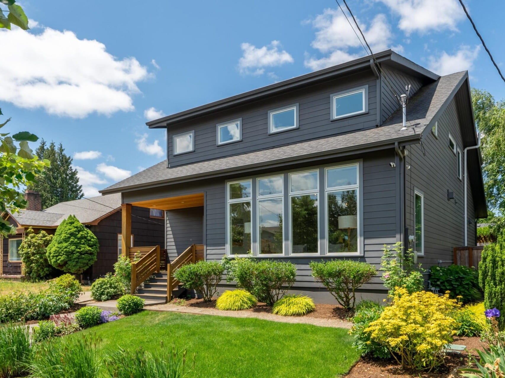 A modern two-story gray house with large windows and a wooden entryway, surrounded by neatly trimmed bushes and colorful flowers. A lush green lawn extends in front under a partly cloudy blue sky.