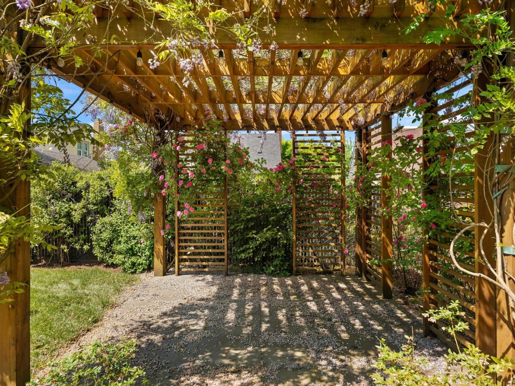 A wooden pergola with a slatted roof and climbing plants creating dappled shade. Pink flowers hang gracefully from the beams. Sunlight filters through, casting shadows on a stone pathway surrounded by lush greenery.