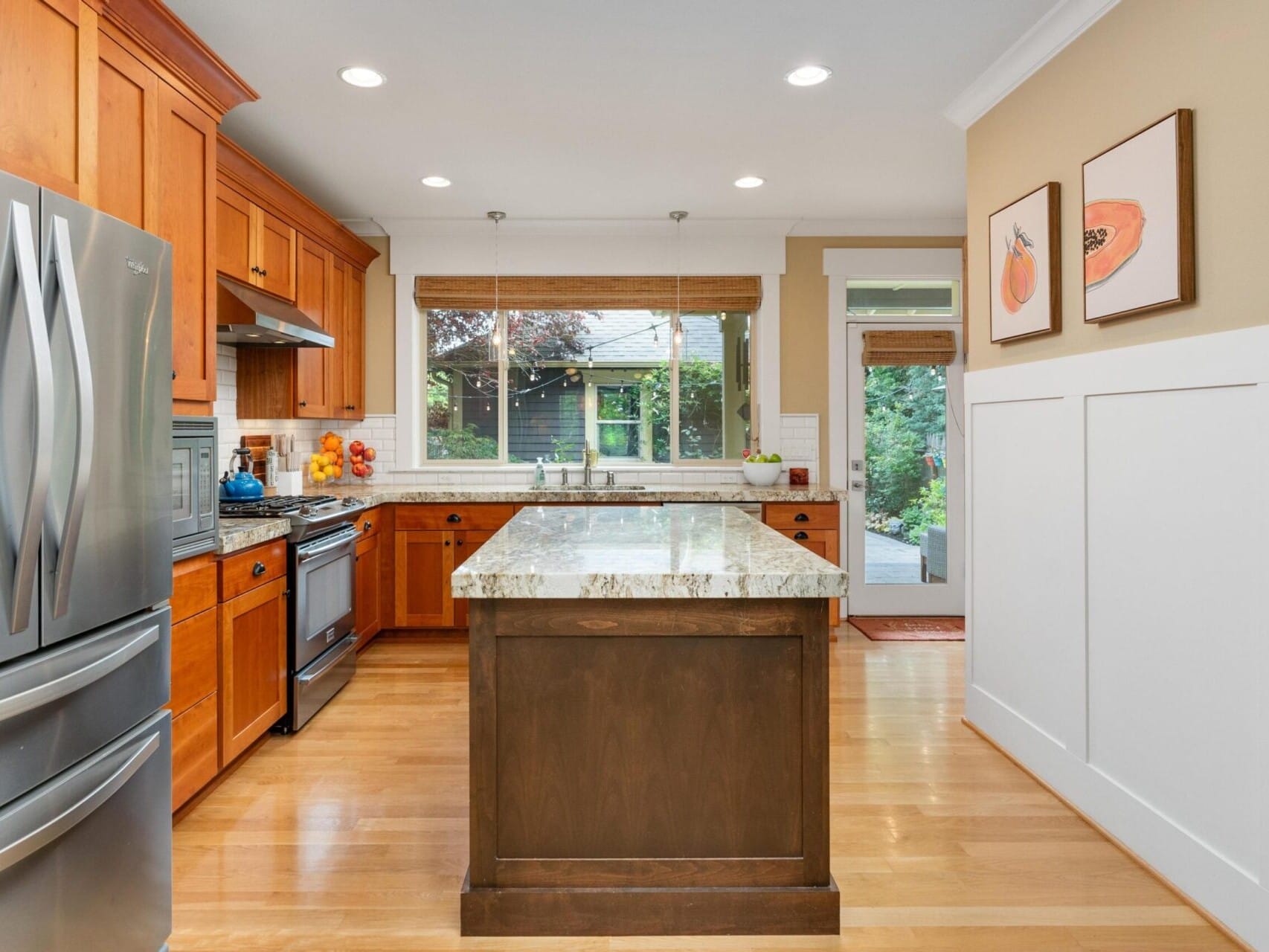 A modern kitchen with wooden cabinets, granite countertops, and stainless steel appliances. A large window offers a view of a garden. Two paintings hang on the beige walls, and the hardwood floor complements the warm tones—truly a gem in Portland Oregon real estate.