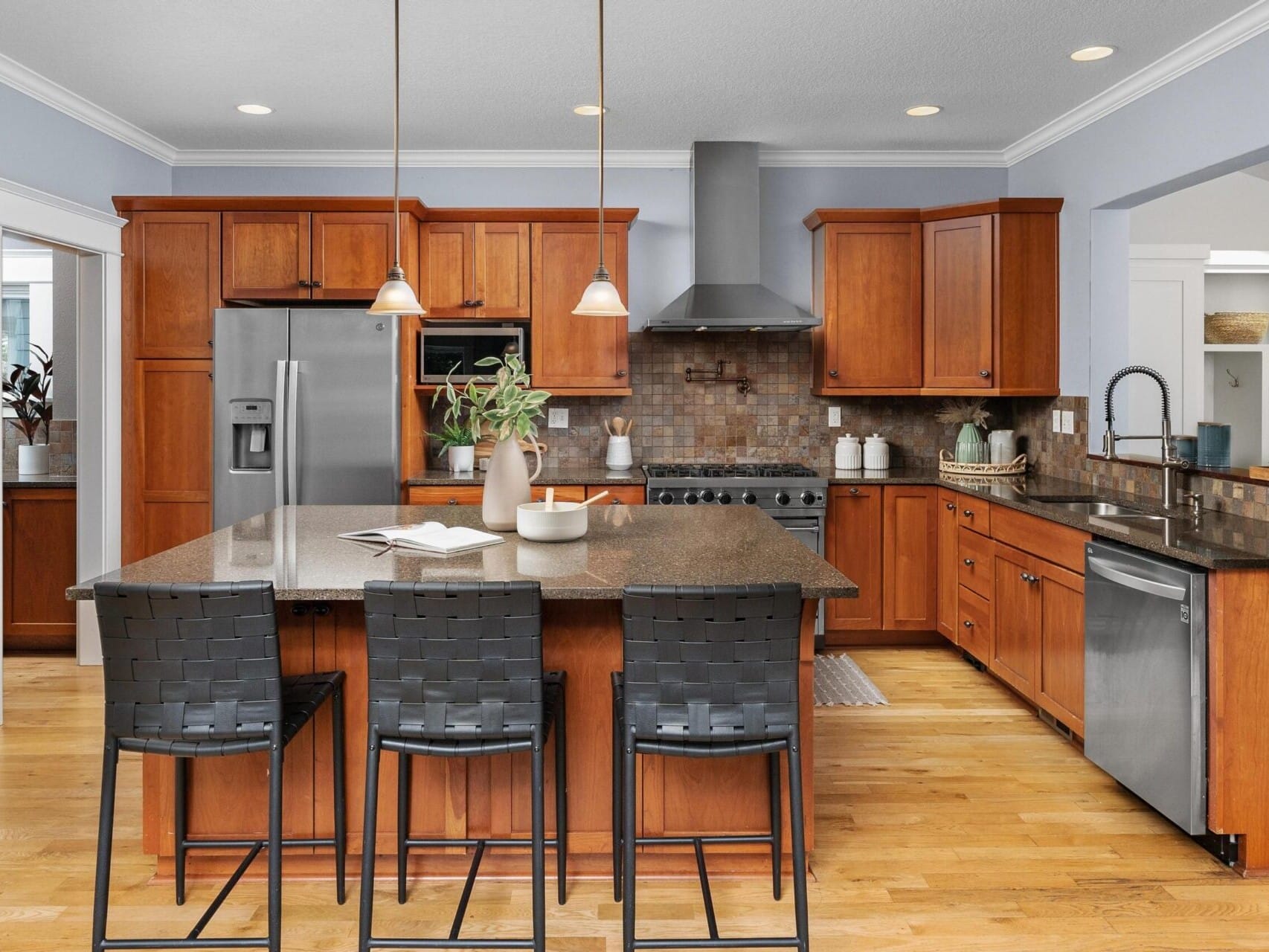 A modern kitchen in Portland real estate boasts wooden cabinets and stainless steel appliances. A large island with a dark countertop is surrounded by three woven barstools. Pendant lights hang above, and plants add a touch of greenery, creating an inviting space for any Portland realtor to showcase.