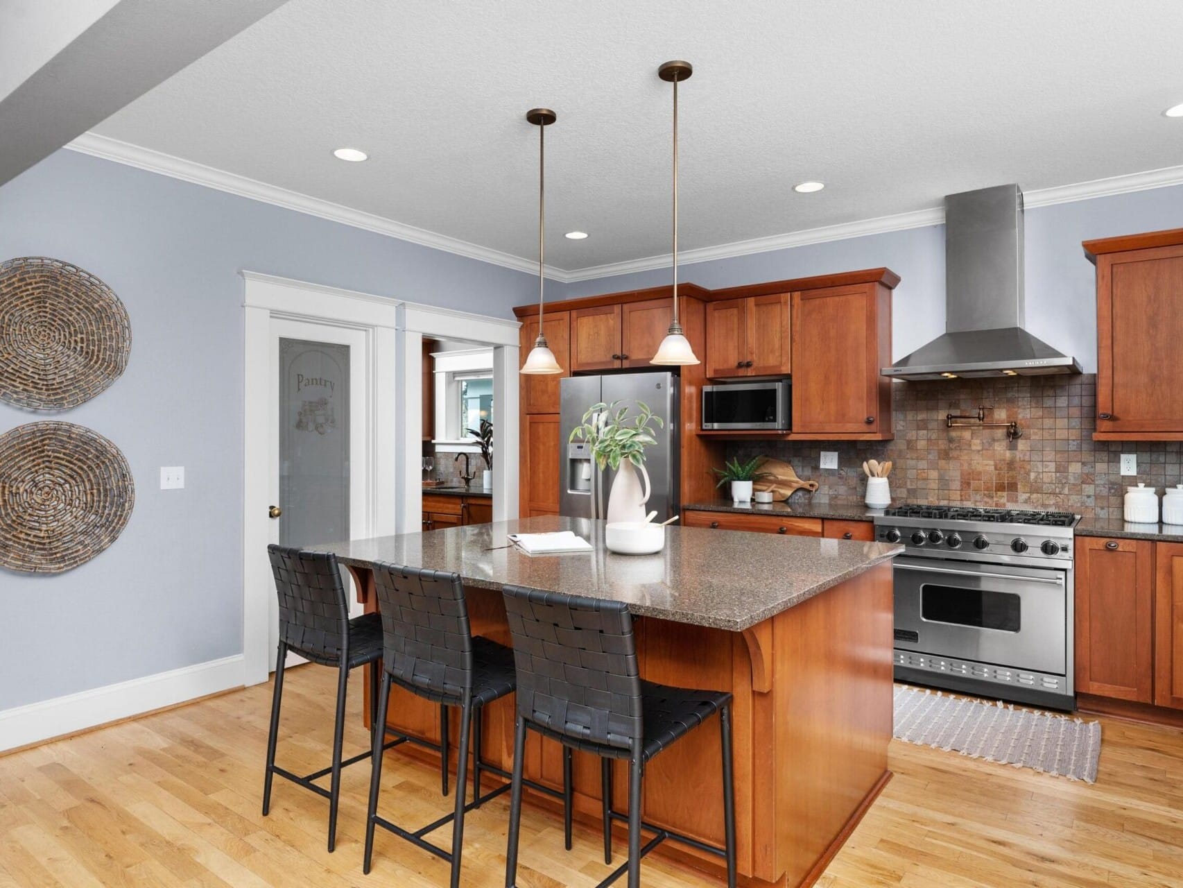 A modern kitchen in a Portland Oregon real estate gem features wooden cabinets and a large island with a dark granite countertop and two woven bar stools. Pendant lights hang above, while stainless steel appliances like the stove and refrigerator shine, complemented by tasteful wall art.