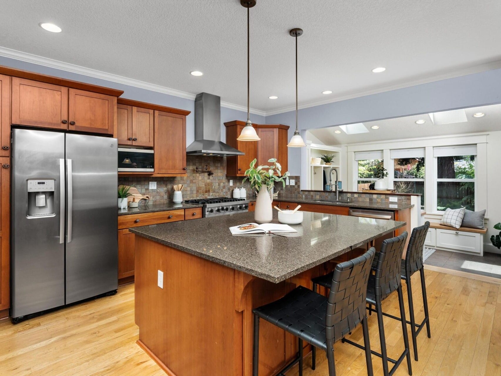 A modern kitchen with wooden cabinets, stainless steel appliances, and a granite island with seating. Bright overhead lights illuminate the space, and large windows offer a view of lush greenery typical of Portland Oregon real estate. A cookbook and plant sit on the island.