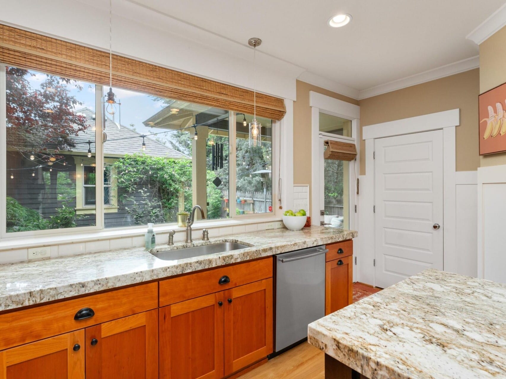 A bright kitchen in a desirable Portland, Oregon real estate listing features large windows, wooden cabinets, and granite countertops. The stainless steel sink and dishwasher sit below the window, with a bowl of green apples on the counter. A door and painting complete this charming space.