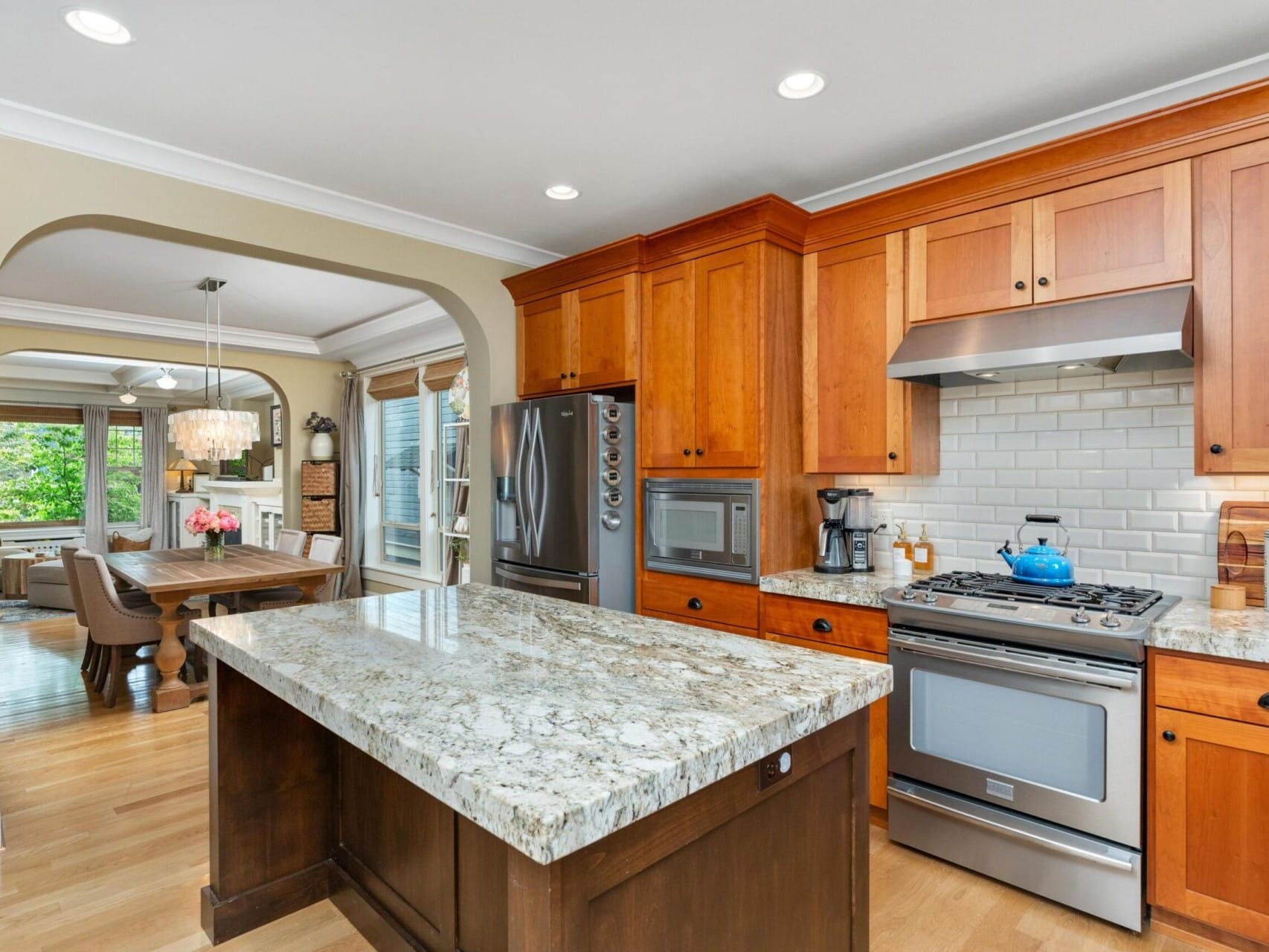 A modern kitchen with wooden cabinets, a stainless steel fridge, and a granite-topped island. A gas stove with a blue kettle sits next to a tile backsplash. An arched opening leads to a dining room with chandelier. With natural light filling the space, it's perfect for any Portland Oregon Real Estate listing.