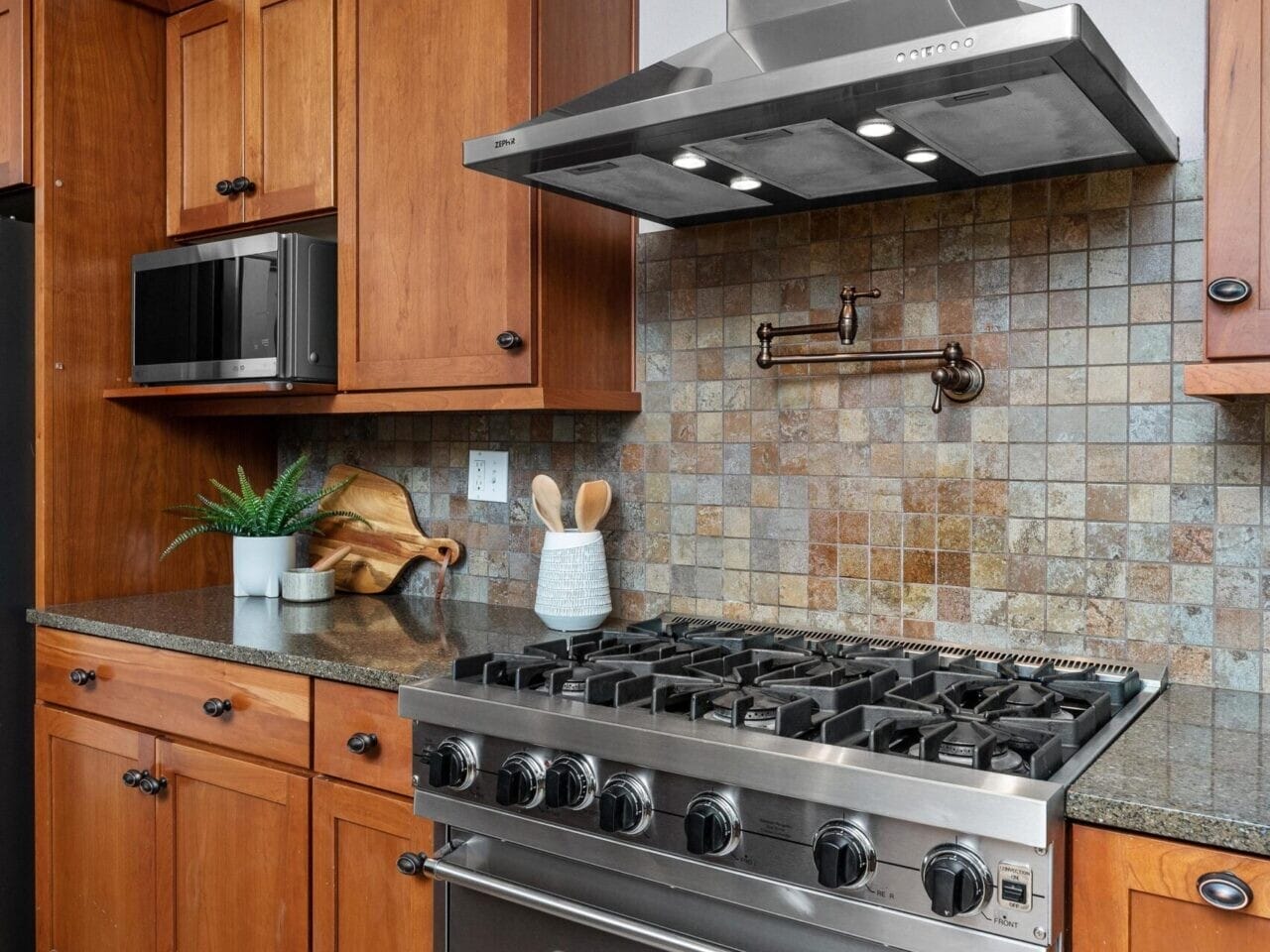 Modern Portland kitchen with wooden cabinets, a stainless steel oven, and a range hood. A tiled backsplash adds texture. Neatly arranged utensils and a potted plant sit on the countertop beside a cutting board. A rug complements the wooden floor, ideal for Portland Oregon real estate seekers.