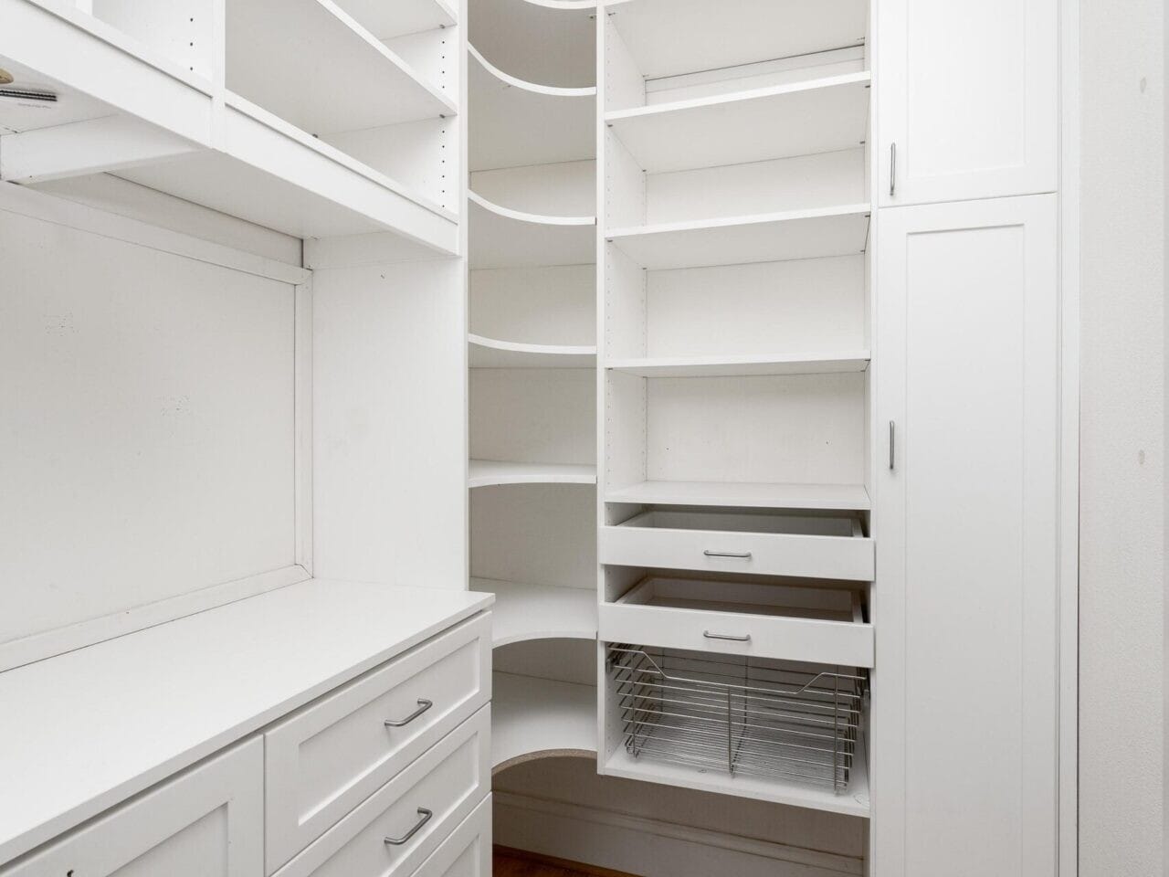 This elegant white walk-in closet boasts open shelves and cabinets with sleek drawers and a long counter on the left. On the right, curved shelves complement a tall cabinet, all set against polished wood floors. Perfect for anyone exploring Portland Oregon real estate for stylish storage solutions!