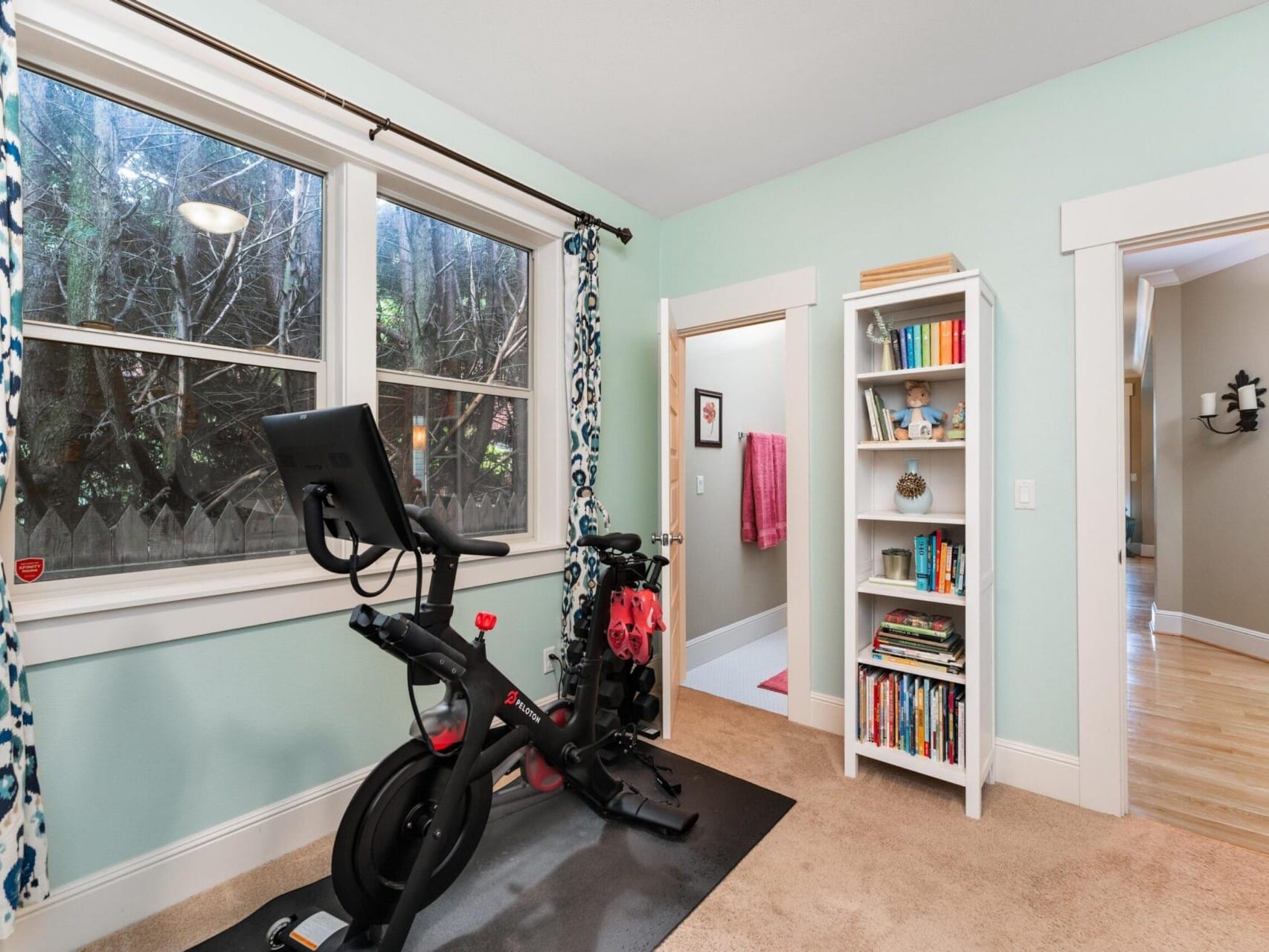 A home exercise room with a stationary bike on a mat near large windows adorned with blue patterned curtains, ideal for those seeking Portland Oregon real estate. A white bookshelf brimming with books and decor complements the light green walls and beige carpet. A hallway and bathroom are visible through a door.