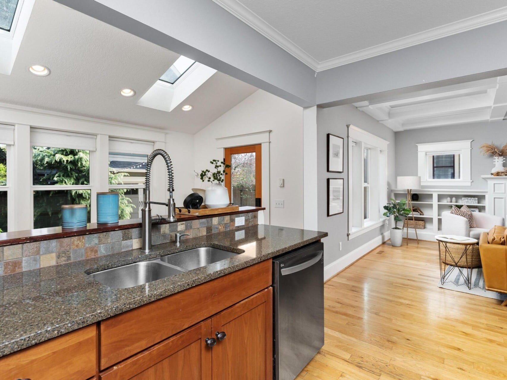 Modern kitchen with a granite countertop and double sink, opening to a bright living room. Skylights, large windows, and light wood flooring create an airy feel. A brown couch and white shelves are visible in the living area—a gem in Portland Oregon real estate.