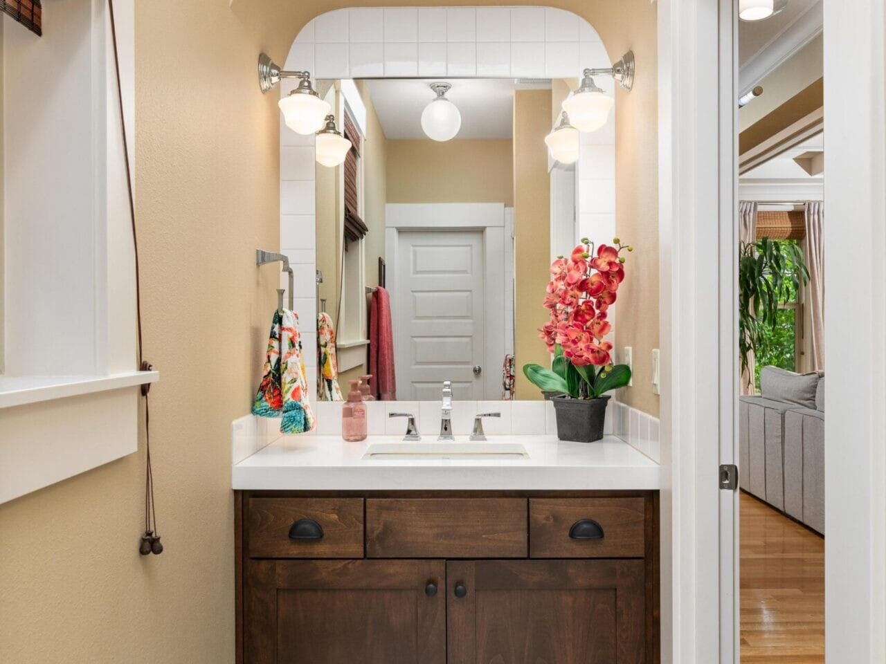 A narrow bathroom with beige walls features a dark wood vanity and white countertop. There's a sink, mirror, and floral decorations. Brightly lit, the space opens to a living area with wooden flooring—perfect for those seeking elegance in Portland Oregon real estate.