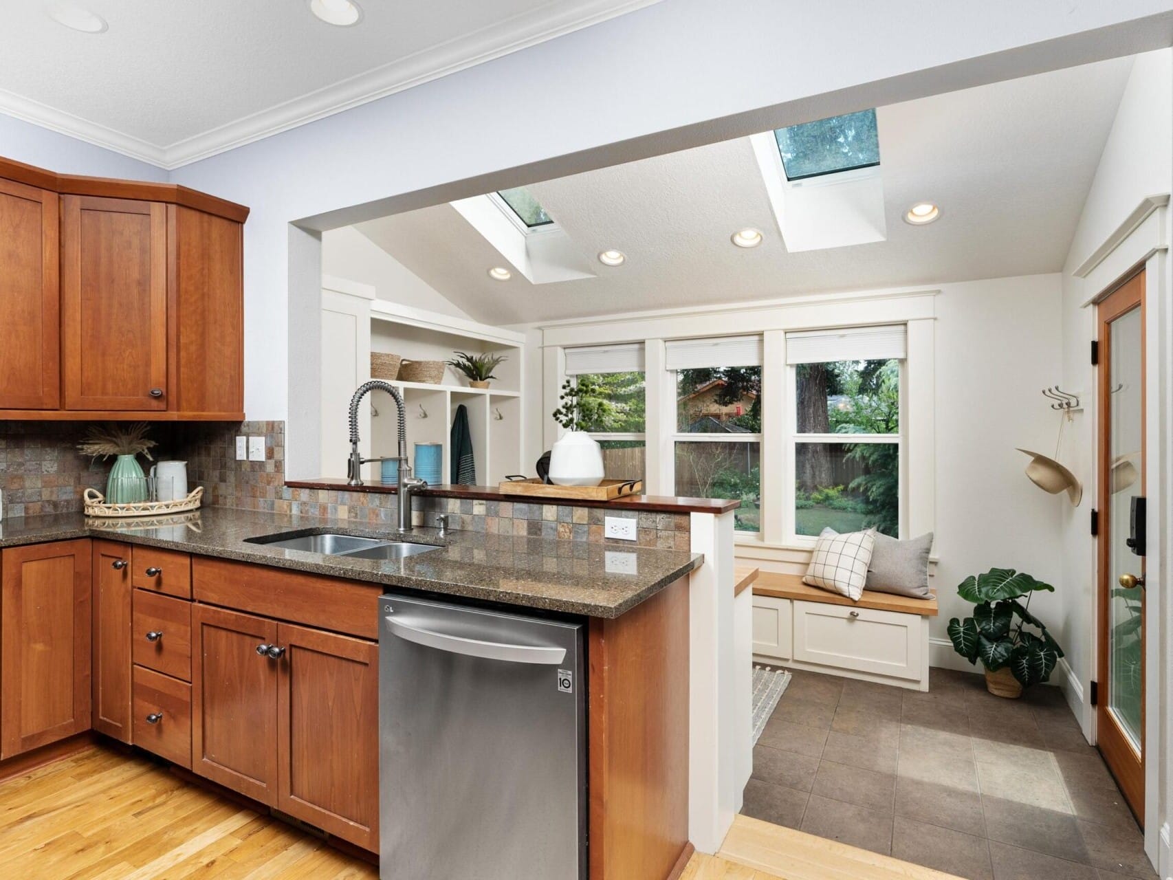 A modern kitchen with brown wooden cabinets, a sleek granite countertop, and a stainless steel dishwasher. Skylights illuminate the adjacent room showcasing Portland Oregon real estate charm with large windows, a cozy bench with cushions, plants, and a hat on the wall.