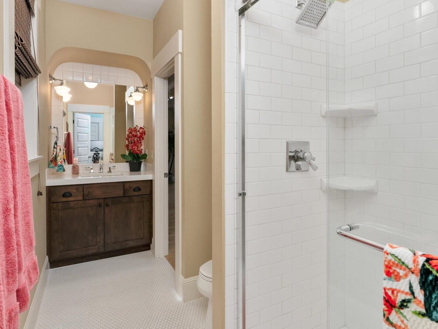 Discover this elegantly designed bathroom, a gem in the Portland Oregon real estate market. It features a glass shower enclosure, white subway tiles, pink towels, and a vanity with a large mirror adorned by red flowers. The charming hexagonal floor tiles complete this top real estate agent's must-see listing.