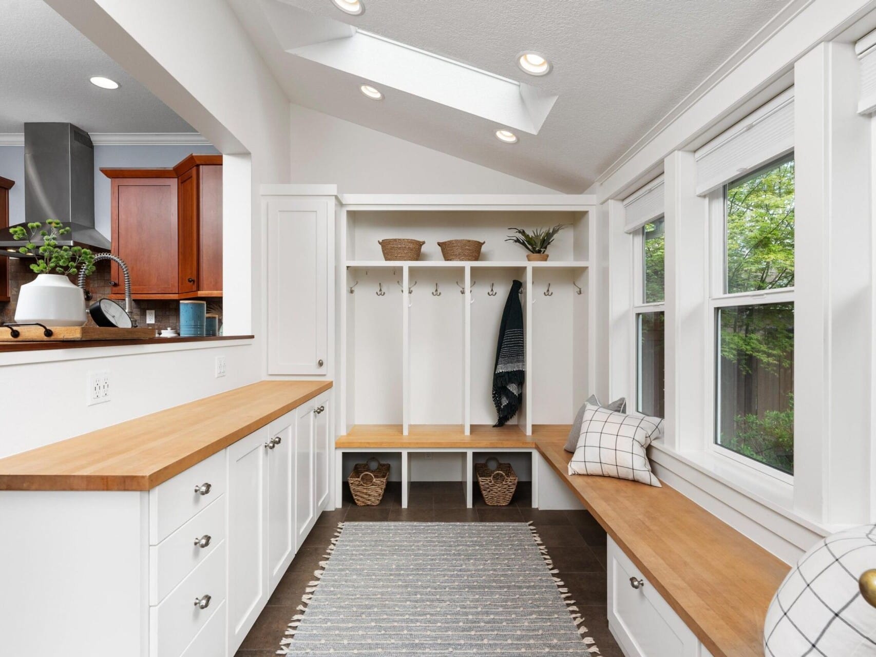 A bright and airy mudroom with white walls and wooden countertops, perfect for Portland Oregon real estate enthusiasts. It features a built-in bench with cubbies and hooks, a striped rug, and large windows letting in natural light. Small decorative plants sit on the counter and shelves.