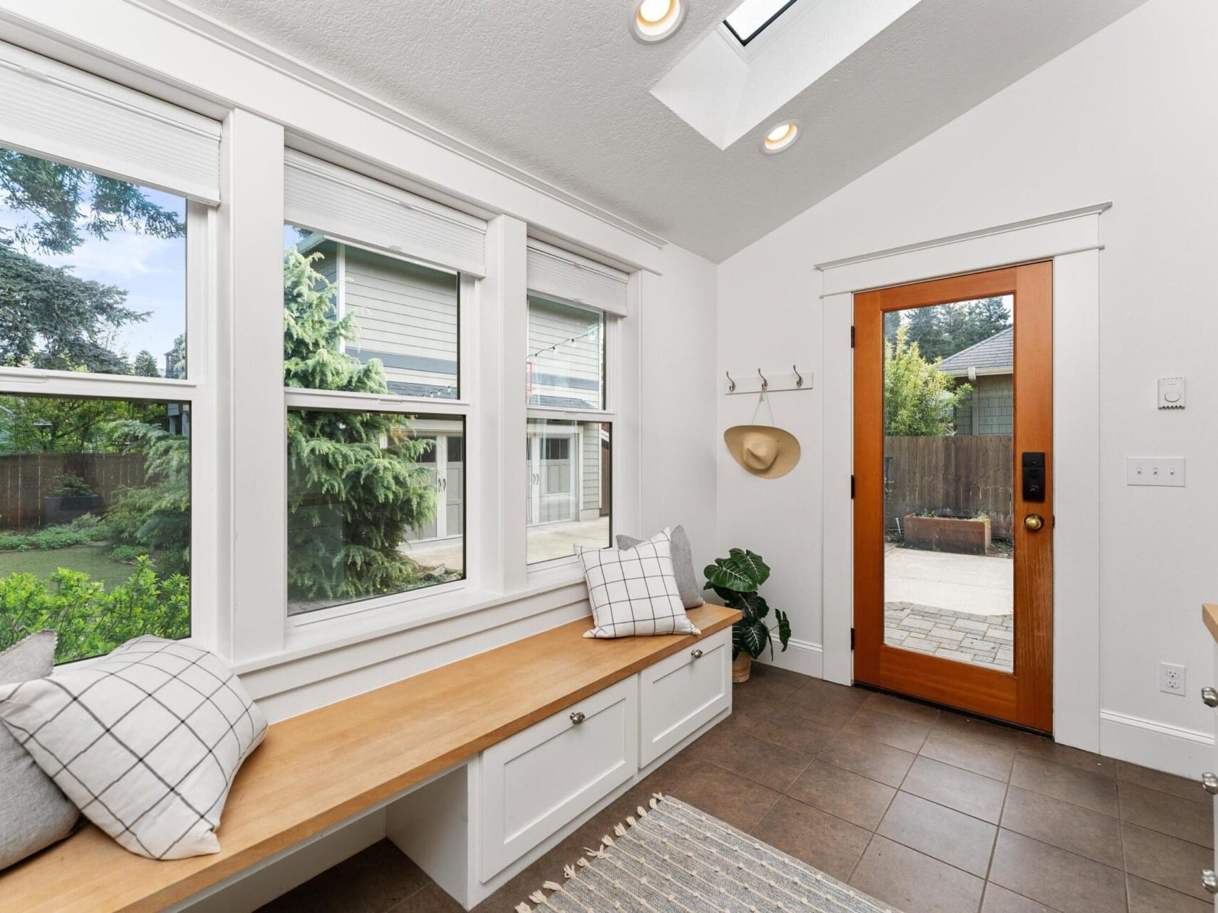 A bright mudroom in a charming Portland, Oregon home features a wooden bench, plaid cushions, and a cozy rug. Windows and a glass door with a wooden frame provide garden views. Wall-mounted hooks and a potted plant add functionality and style, while skylights illuminate the space.