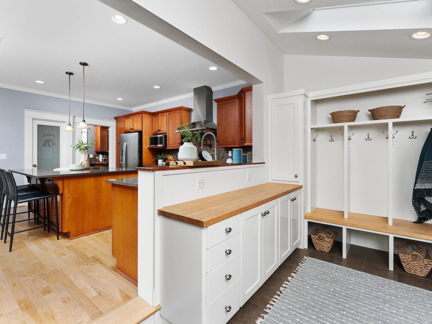 A modern kitchen in Portland, Oregon real estate features wooden cabinets, stainless steel appliances, and an island with bar stools. An adjacent mudroom area boasts white storage cubbies, shelves with baskets, and a blue rug on dark flooring—a perfect find with a top real estate agent.