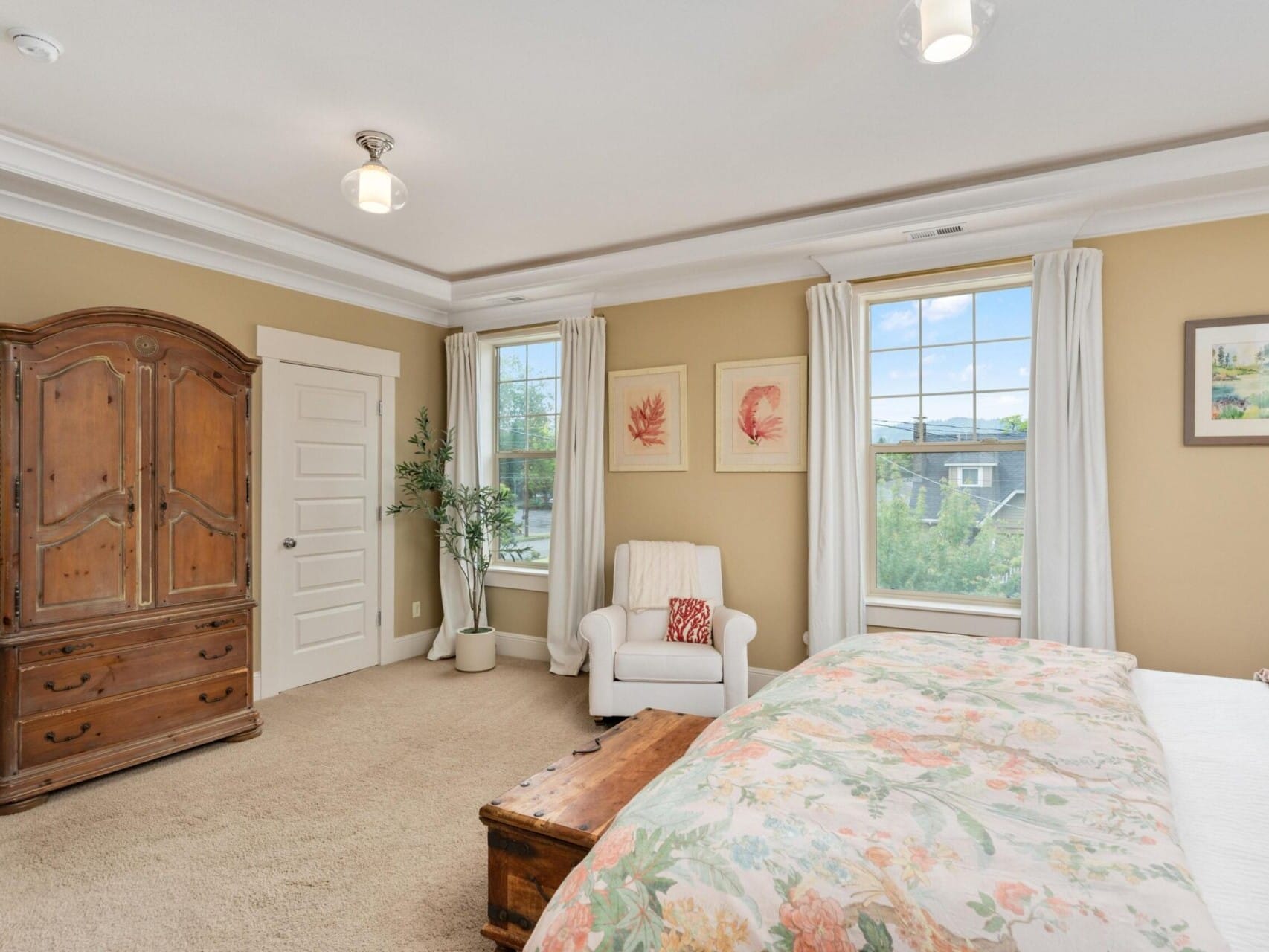 A cozy bedroom in Portland, Oregon real estate, featuring a large wooden wardrobe, an armchair, and a bed with a floral-patterned comforter. The room has beige walls, two windows with white curtains, a small potted plant, and framed artwork—a gem any top Portland realtor would love to show.