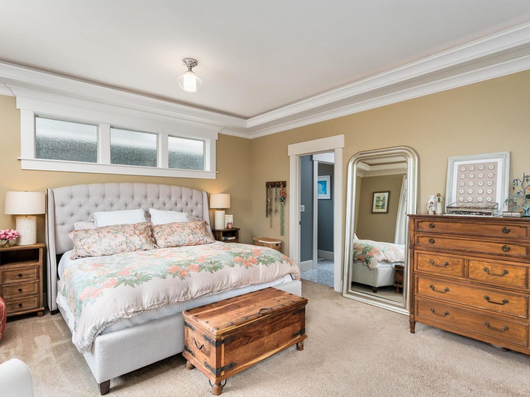 In a slice of Portland Oregon real estate, this cozy bedroom boasts a tufted bed with floral bedding, two wooden nightstands with lamps, a large ornate mirror, and a vintage dresser. Soft lighting and neutral walls create a warm atmosphere. A wooden chest sits at the foot of the bed.