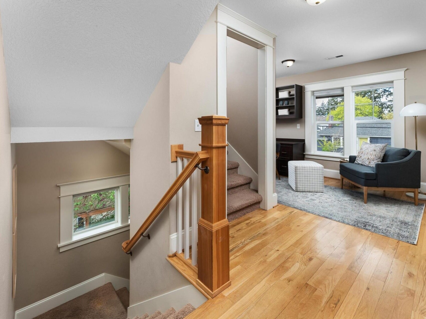 At the top of a wooden staircase, this cozy room features a dark sofa, an ottoman, a floor lamp, and a small desk. A window bathes the space in natural light, highlighting warm wooden floors. This charming setting is perfect for those exploring Portland Oregon real estate opportunities.